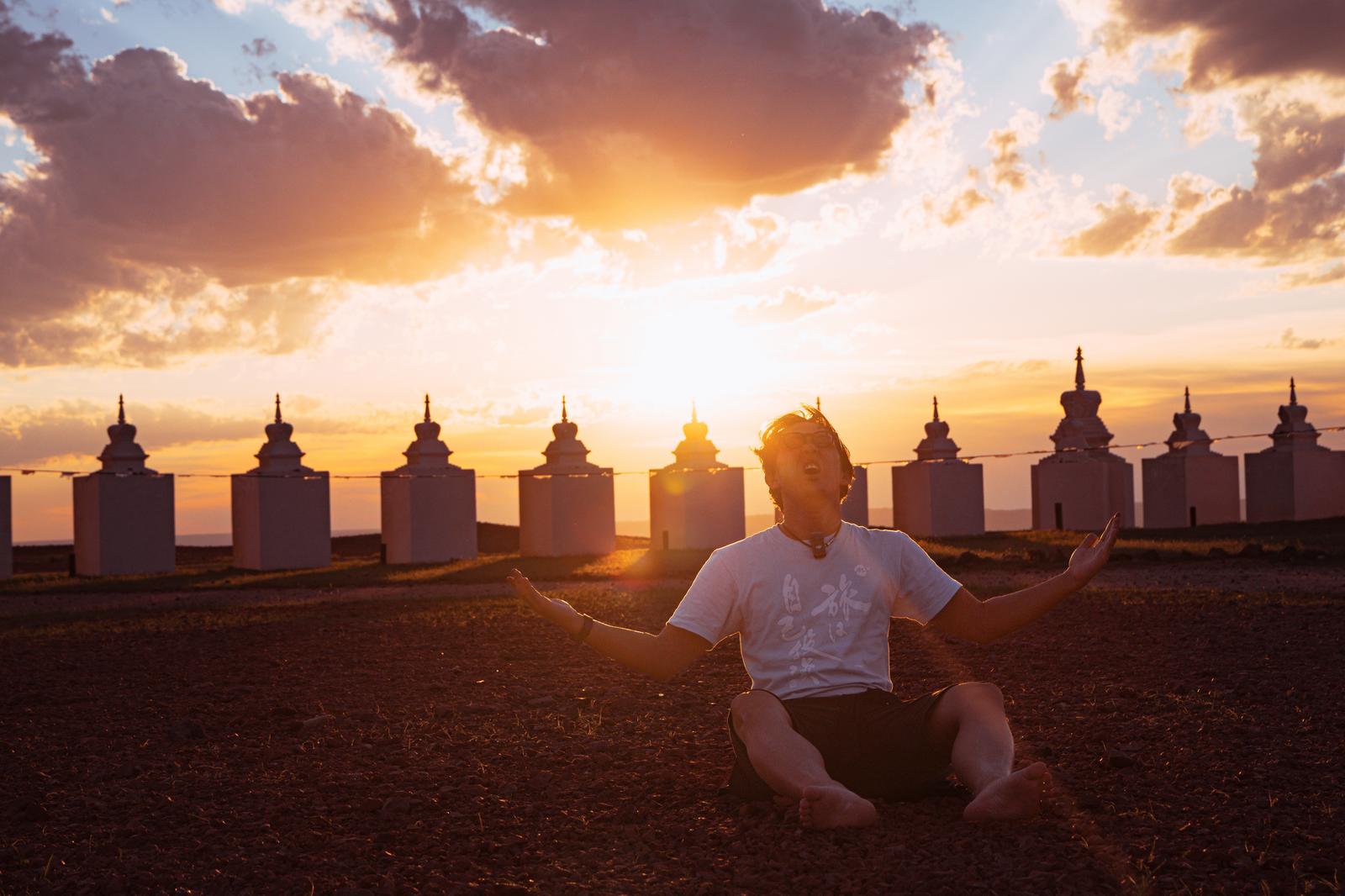 夕焼けを背景に白い仏塔が並ぶモンゴルで、両腕を広げて座る白Tシャツの男性［モデル：もーりー］